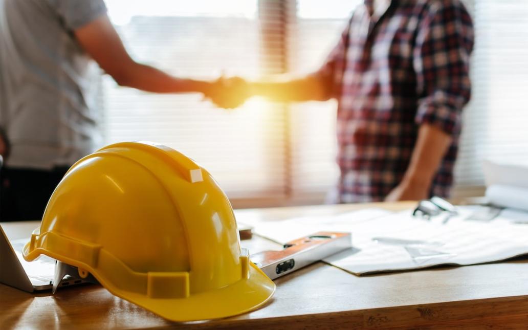 Two construction workers shake hands behind a table that has documents and a yellow hard hat on it