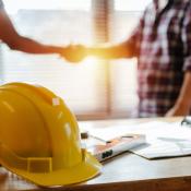 Two construction workers shake hands behind a table that has documents and a yellow hard hat on it