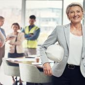 Woman in blazer smiles and holds hard hat, behind her are four construction workers conversing
