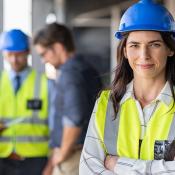 Woman in blue hard hat and yellow construction stands in foreground, while three men stand together in background looking over documents